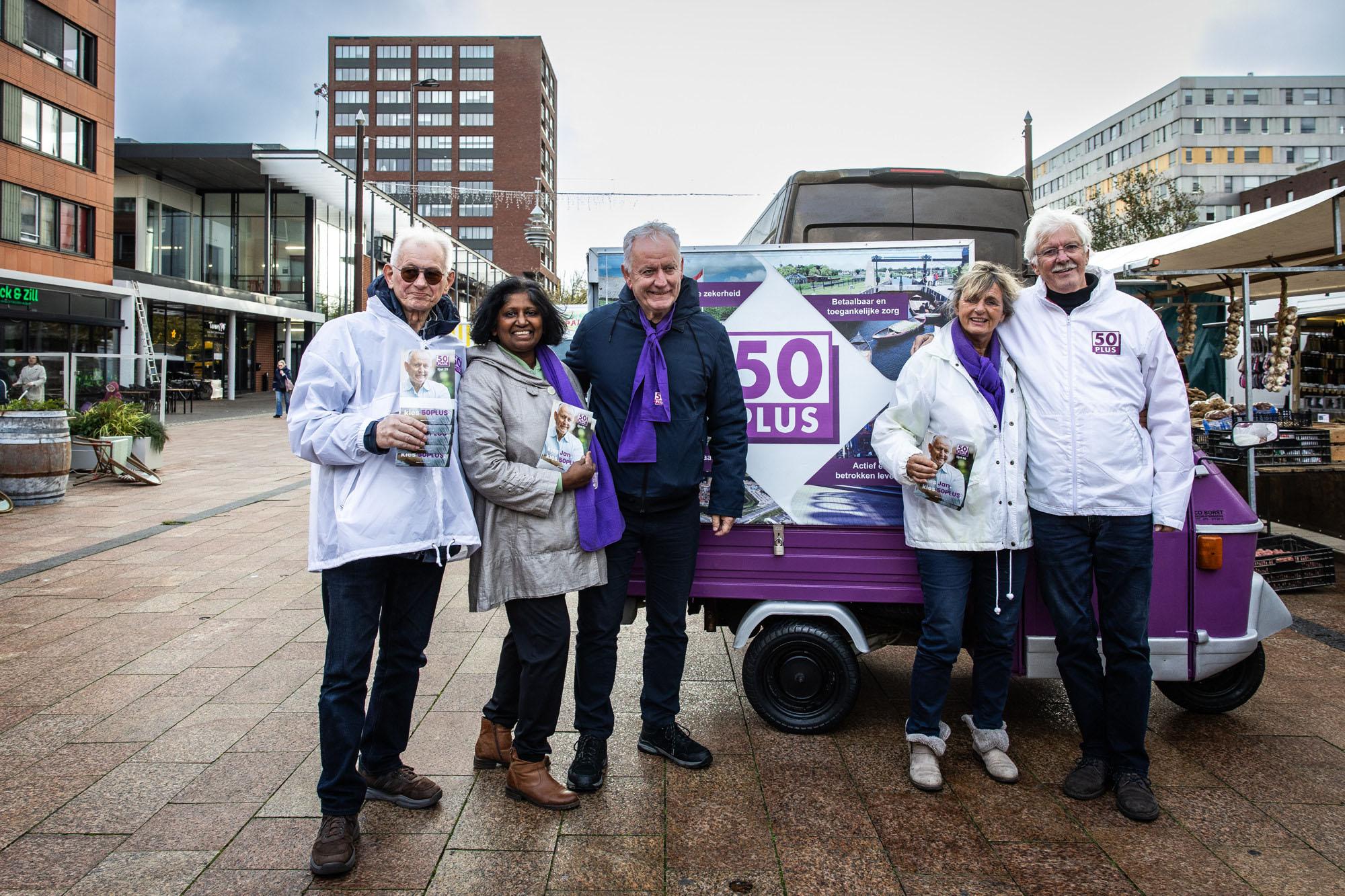 Een foto van lijsttrekker Jan Struijs van de partij 50Plus, samen met andere campagnevoerders, op een plein. Ze dragen 50Plus-kleding en achter hen staat een campagnebusje van de partij.