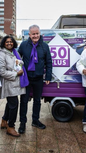 Een foto van lijsttrekker Jan Struijs van de partij 50Plus, samen met andere campagnevoerders, op een plein. Ze dragen 50Plus-kleding en achter hen staat een campagnebusje van de partij.