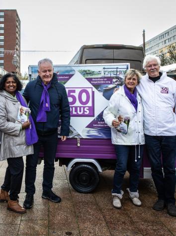 Een foto van lijsttrekker Jan Struijs van de partij 50Plus, samen met andere campagnevoerders, op een plein. Ze dragen 50Plus-kleding en achter hen staat een campagnebusje van de partij.