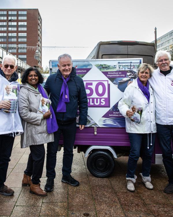 Een foto van lijsttrekker Jan Struijs van de partij 50Plus, samen met andere campagnevoerders, op een plein. Ze dragen 50Plus-kleding en achter hen staat een campagnebusje van de partij.