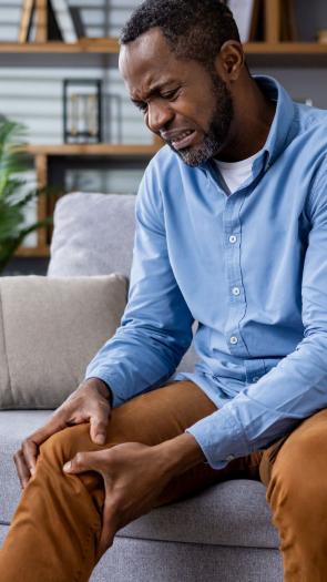 Een foto van een man die intense pijn heeft in zijn been en daar met zijn handen heen grijpt. Hij zit op een bank in zijn woonkamer.