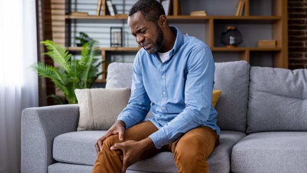 Een foto van een man die intense pijn heeft in zijn been en daar met zijn handen heen grijpt. Hij zit op een bank in zijn woonkamer.