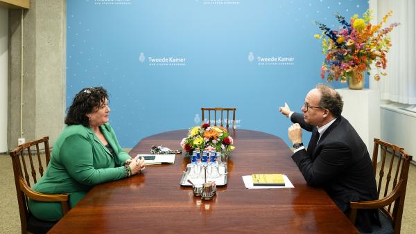 Verkenner Koolmees zit met Caroline van der Plas aan houten tafel in gesprek bij muur waar Tweede Kamer op staat