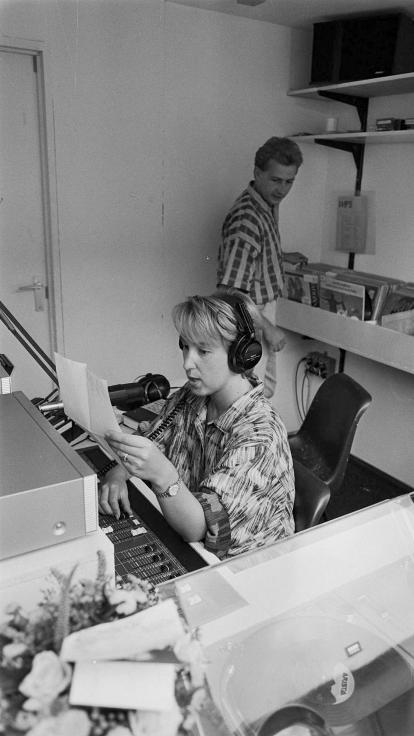 Een zwart-witfoto van twee radiopiraten die radio maken. Zij zit op de voorgrond en leest iets voor in de microfoon. Hij is op de achtergrond door lp-platen aan het zoeken.