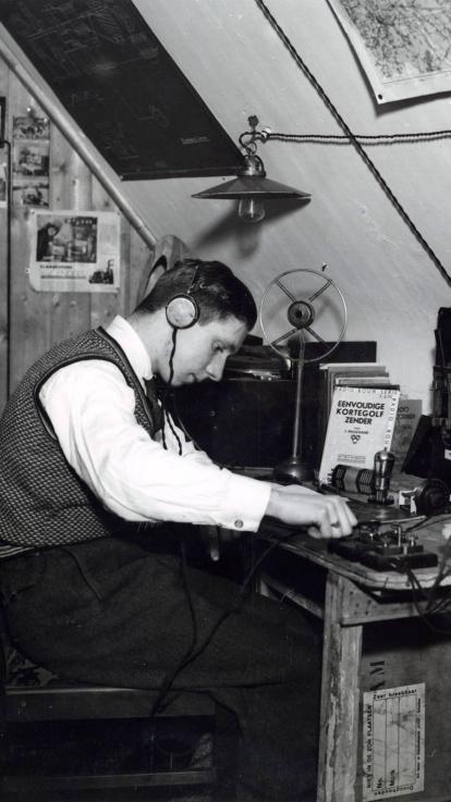 Een zwart-witfoto van een zendamateur uit de jaren 30 die op zolder aan zijn zelfgemaakte radioapparatuur sleutelt.