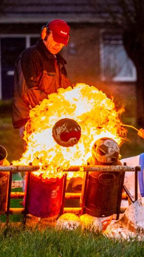 Twee mannen steken met een gasbrander melkbussen met carbid af, terwijl een grote vlam en een weggeschoten voetbal te zien zijn. 