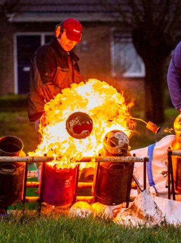 Twee mannen steken met een gasbrander melkbussen met carbid af, terwijl een grote vlam en een weggeschoten voetbal te zien zijn.