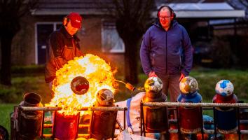 Twee mannen steken met een gasbrander melkbussen met carbid af, terwijl een grote vlam en een weggeschoten voetbal te zien zijn. 
