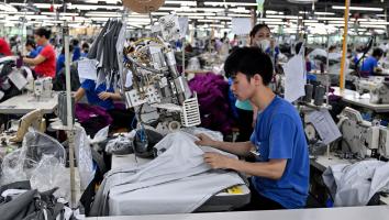 Grote zaal waar veel mensen achter naaimachines zitten. Een jonge jongen is de focus in het midden