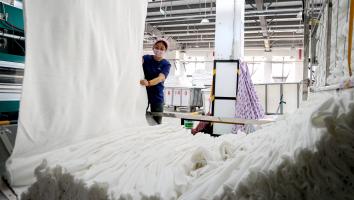 Een vrouw met mondkapje houdt enorme stukken stof vast terwijl die uit een machine komen