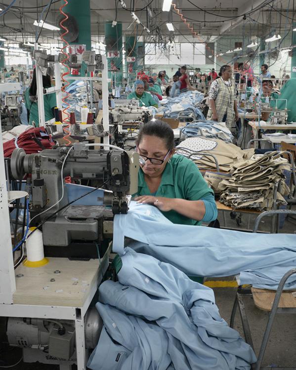 Vrouw zit in een grote zaal achter een naaimachine te werken. Om haar heen zitten overal ook mensen achter naaimachines.