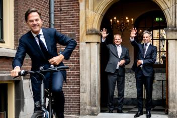 Een foto van premier Rutte die wegfietst bij het Torentje, en Schoof en iemand anders die hem uitzwaaien vanaf de boog bij de uitgang.