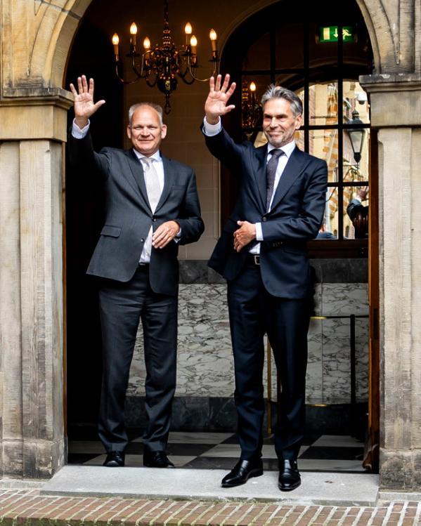 Een foto van premier Rutte die wegfietst bij het Torentje, en Schoof en iemand anders die hem uitzwaaien vanaf de boog bij de uitgang.