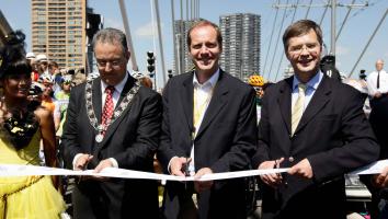 Drie bestuurders, waaronder premier Balkenende, openen een evenement door gezamenlijk een lint door te knippen, omringd door toeschouwers en wielrenners.