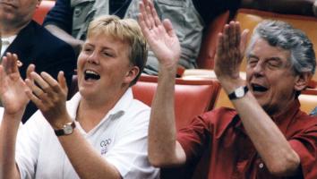 Oud-premier Wim Kok en prins Willem-Alexander klappen enthousiast op de tribune tijdens een sportwedstrijd, samen met andere toeschouwers.
