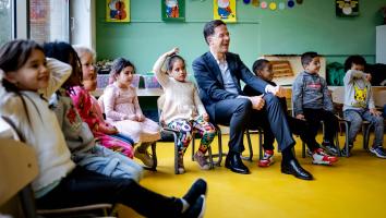 Mark Rutte zit lachend op een stoeltje in de kring van een kleuterklas op een basisschool tussen de leerlingen.