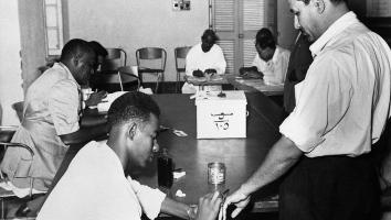 Zwart-wit foto van een man met een sigaret in zijn mond die een stempel op zijn hand krijgt van een man die aan tafel zit. Op het midden van de tafel staat een witte stemdoos. 