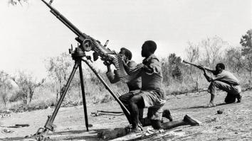 Zwart-wit foto van 3 mannen in legeruniform hurkend bij vuurwapens die op driepoten staan. 