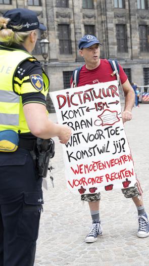 Demonstrant met protestbord met de tekst: Dictatuur komt eraan. Wat voor toekomst wil jij? met twee agenten in uniform om hem heen. 