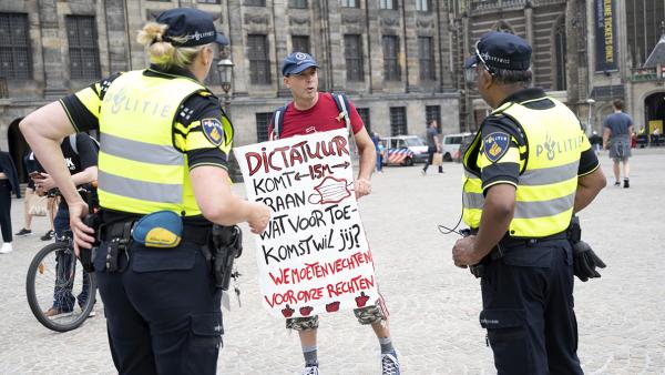 Demonstrant met protestbord met de tekst: Dictatuur komt eraan. Wat voor toekomst wil jij? met twee agenten in uniform om hem heen. 