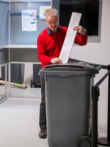 Man stopt stembiljet in een kliko terwijl op de achtergrond een vrouw in een hokje aan het stemmen is