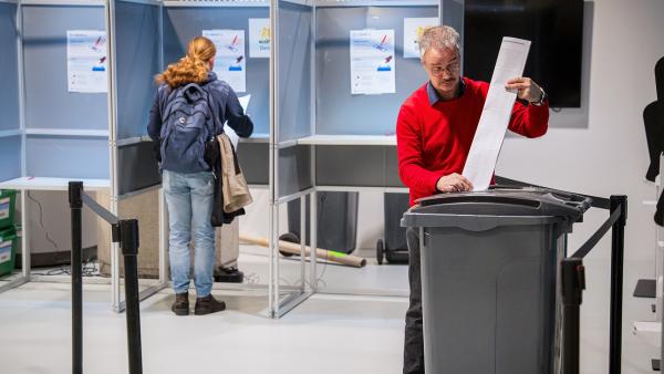 Man stopt stembiljet in een kliko terwijl op de achtergrond een vrouw in een hokje aan het stemmen is