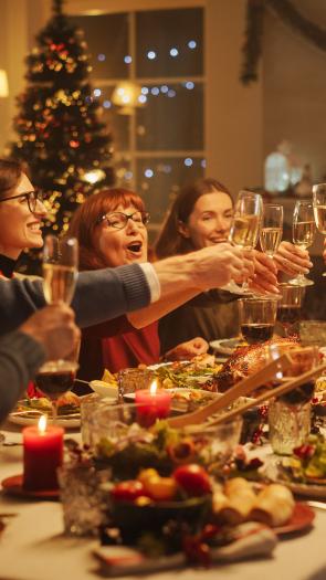 Een foto van volwassenen die aan tafel proosten met Kerst met feestversiering op de achtergrond en lekker eten op tafel.