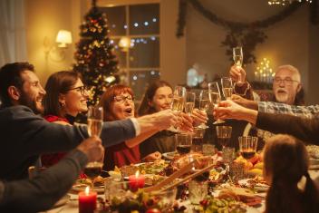 Een foto van volwassenen die aan tafel proosten met Kerst met feestversiering op de achtergrond en lekker eten op tafel.