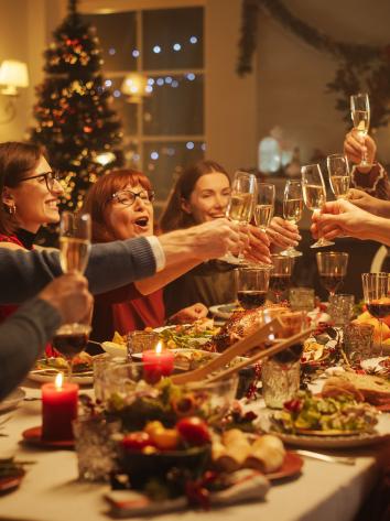 Een foto van volwassenen die aan tafel proosten met Kerst met feestversiering op de achtergrond en lekker eten op tafel.