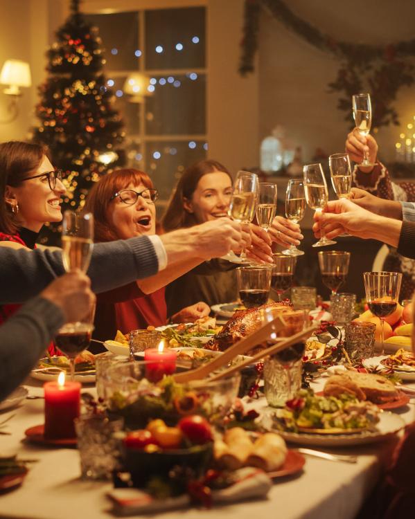 Een foto van volwassenen die aan tafel proosten met Kerst met feestversiering op de achtergrond en lekker eten op tafel.