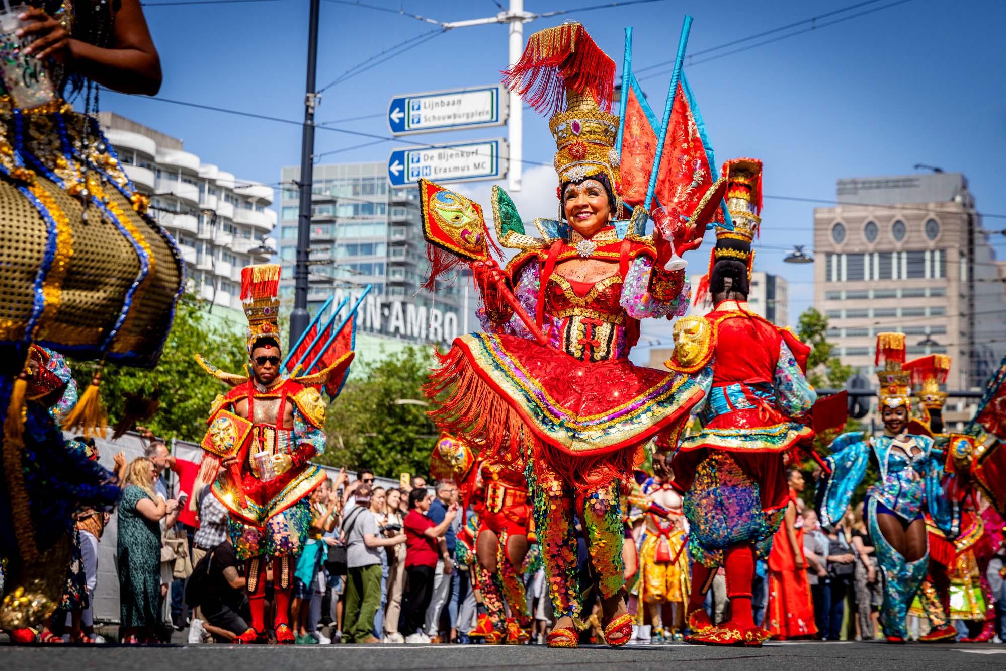 Een foto van de viering van Zomercarnaval in Rotterdam. Vooraan in de optocht een vrouw in feestelijke, kleurrijke kleding.