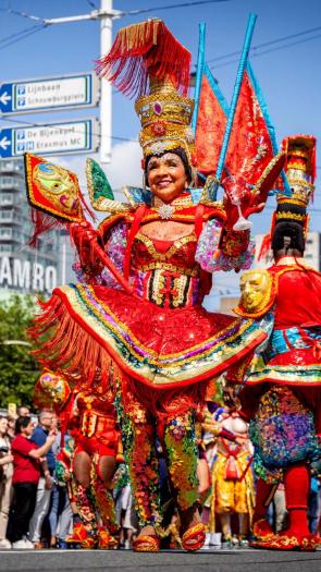 Een foto van de viering van Zomercarnaval in Rotterdam. Vooraan in de optocht een vrouw in feestelijke, kleurrijke kleding.