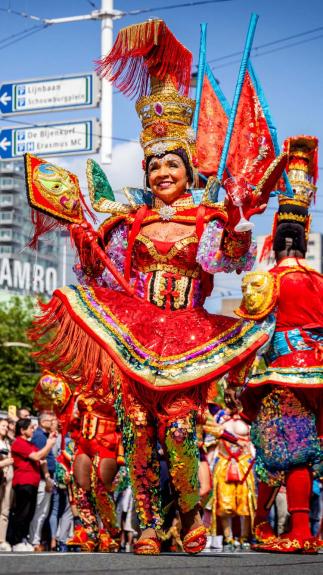 Een foto van de viering van Zomercarnaval in Rotterdam. Vooraan in de optocht een vrouw in feestelijke, kleurrijke kleding.