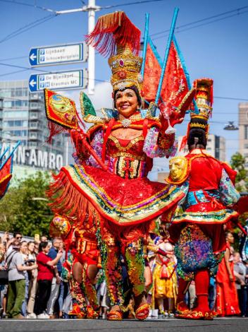 Een foto van de viering van Zomercarnaval in Rotterdam. Vooraan in de optocht een vrouw in feestelijke, kleurrijke kleding.
