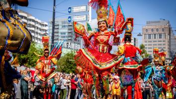 Een foto van de viering van Zomercarnaval in Rotterdam. Vooraan in de optocht een vrouw in feestelijke, kleurrijke kleding.