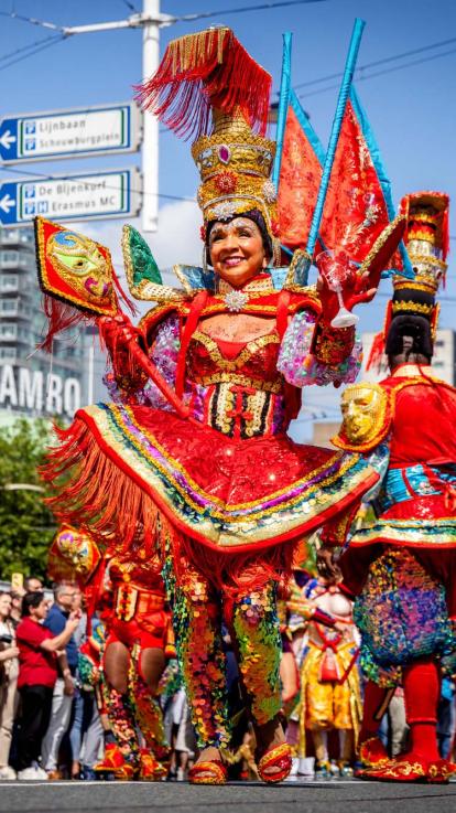 Een foto van de viering van Zomercarnaval in Rotterdam. Vooraan in de optocht een vrouw in feestelijke, kleurrijke kleding.