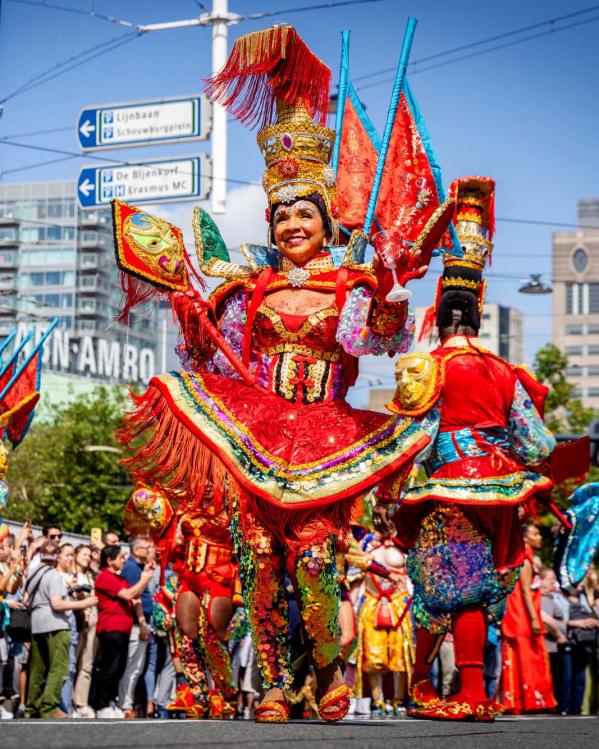 Een foto van de viering van Zomercarnaval in Rotterdam. Vooraan in de optocht een vrouw in feestelijke, kleurrijke kleding.