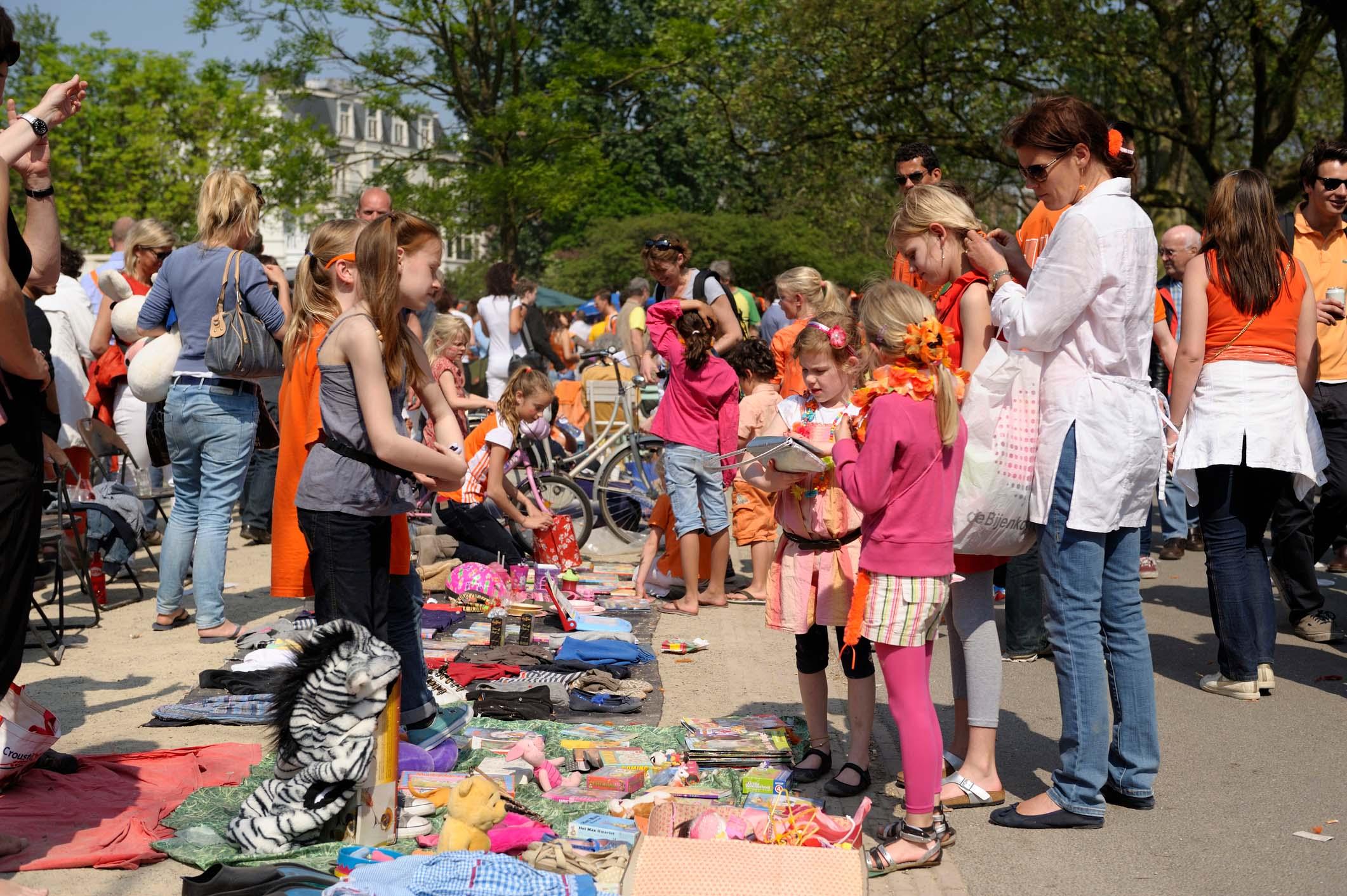 Een vrijmarkt op een plein tijdens Koningsdag. Er liggen kleden op de grond met allerlei spullen en veel mensen lopen rond.