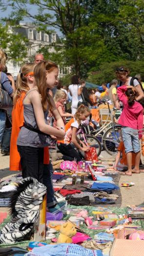 Een vrijmarkt op een plein tijdens Koningsdag. Er liggen kleden op de grond met allerlei spullen en veel mensen lopen rond.