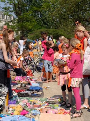 Een vrijmarkt op een plein tijdens Koningsdag. Er liggen kleden op de grond met allerlei spullen en veel mensen lopen rond.