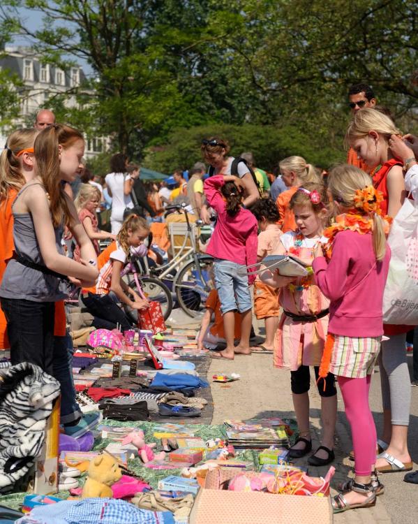 Een vrijmarkt op een plein tijdens Koningsdag. Er liggen kleden op de grond met allerlei spullen en veel mensen lopen rond.