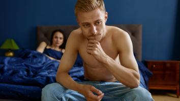 Een man zit half ontbloot op de rand van het bed en kijkt bedenkelijk voor zich uit. In het bed ligt een vrouw die naar hem kijkt. 