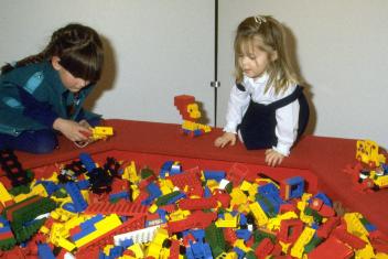 Twee meisjes spelen met lego in de jaren 80, voor hen een grote bak met felgekleurde legostenen.