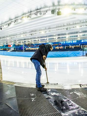 Een ijsmeester maakt het ijs klaar bij de schaatsbaan in Thialf, op de achtergrond zie je de schaatsbanen en lege zaal.