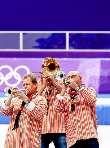 Drie leden van het dweilorkest blazen op hun blaasinstrumenten op de ijsbaan tijdens de Olympische Spelen.