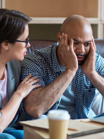 Man kijkt gefrustreerd naar laptop met handen tegen zijn kale hoofd aan. Vrouw naast hem legt hand op schouder om hem gerust te stellen