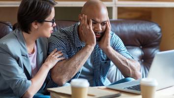 Man kijkt gefrustreerd naar laptop met handen tegen zijn kale hoofd aan. Vrouw naast hem legt hand op schouder om hem gerust te stellen