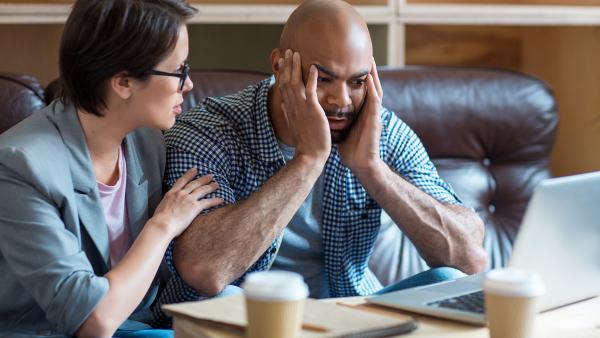 Man kijkt gefrustreerd naar laptop met handen tegen zijn kale hoofd aan. Vrouw naast hem legt hand op schouder om hem gerust te stellen