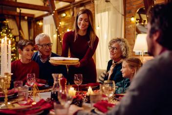 Familie aan het dineren aan een tafel vol kaarsen. Met ouderen en kinderen. Jonge vrouw zet lachend een taart op tafel. 