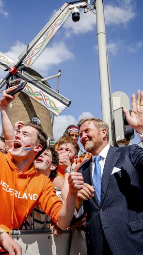 Koning Willem-Alexander maakt een selfie met mensen gekleed in oranje die bij een molen staan achter een hek